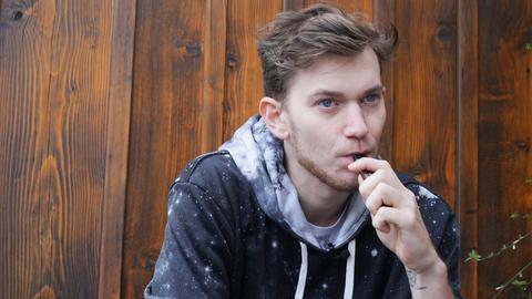 Ein junger Mann lehnt an einer Holzwand und raucht eine e-Zigarette.