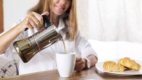 Eine Frau gießt sich am Frühstückstisch mit Croissants eine Tasse Kaffee ein.