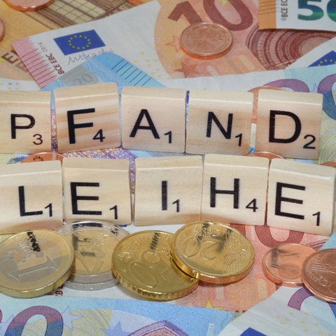 Scrabbleplättchen zeigen das Wort "Pfandleihe", sie liegen auf Geldscheinen. Vor und hinter ihnen liegen mehrere Euromünzen.