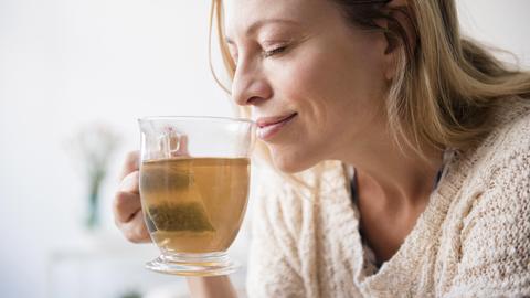 Eine Frau im Strickpullover riecht an einer Tasse Tee und lächelt dabei.