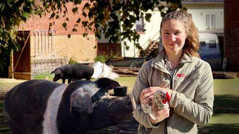 Bäuerin Franziska mit einem Huhn im Arm, im Hintergrund sieht man zwei Schweine. 