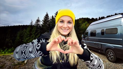 Maike Tschorn bildet mit ihren Fingern ein Herz, im Hintergrund der Campervan. 