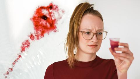 Eine junge Frau hält eine gefüllte Menstruationstasse in der Hand und schaut sie skeptisch an. Im Hintergrund sieht man in Wasser aufgelöstes Blut im Abfluss verschwinden (Collage).