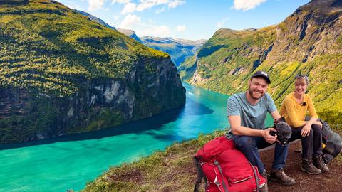 Nina und Jonas sitzen strahlend auf einer Bank mit Blick auf den Geirangerfjord in Norwegen.
