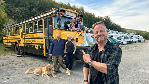 Friso Richter deutet strahlend mit dem Zeigefinger auf den umgebauten US-amerikanischen Schulbus im Hintergrund, vor und auf dem mehrere Personen mit Hund stehen oder sitzen.