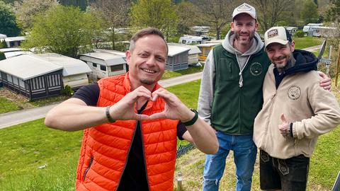 Friso Richter formt mit seinen Händen ein Herz. Er steht neben zwei Männern, die sich in den Armen halten. Im Hintergrund ist ein Campingplatz.