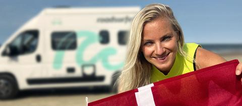 Reporterin Meike hält dänische Flagge in die Kamera, im Hintergrund Strand und Camper.