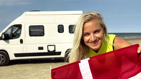 Reporterin Meike hält dänische Flagge in die Kamera, im Hintergrund Strand und Camper.