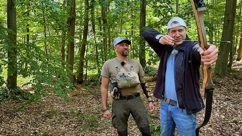 Danny Morgenstern an Pfeil und Bogen. Er steht mit Trainer Justin Kay in einem Wald.