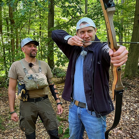 Danny Morgenstern an Pfeil und Bogen. Er steht mit Trainer Justin Kay in einem Wald.