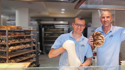 Zwei Protagonisten stehen in einer Bäckerei und blicken freundlich in die Kamera.