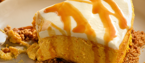 Ein Stückchen Kürbiskuchen mit Frischkäse-Frosting auf einem Teller. Im Hintergrund der angeschnittene Kuchen und mehrere Zierkürbisse.