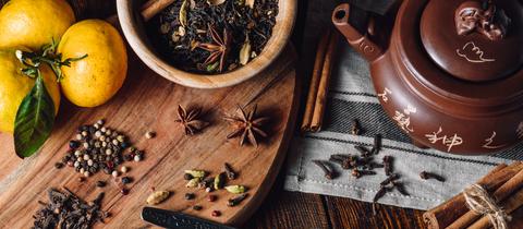 Zutaten für einen Masala Chai: Gewürze wie Anis, Zimt und Pfefferkörner auf Holzgrund. Daneben eine Teekanne.
