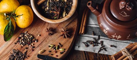 Zutaten für einen Masala Chai: Gewürze wie Anis, Zimt und Pfefferkörner auf Holzgrund. Daneben eine Teekanne.