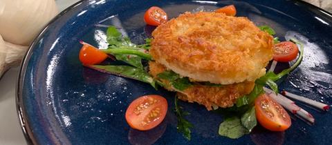 Sellerie-Knusper-Schnitzel mit nussigem Kräutersalat auf dunklem Teller. Im Hintergrund rohes Gemüse wie Knollensellerie, Möhren und Knoblauch.