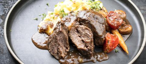 Traditionelle deutsche geschmorte Schweinebacken in brauner Soße mit Champignons und Kartoffelpüree als Nahaufnahme auf einem modernen Designteller