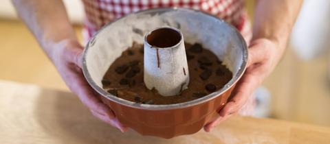 Ein Mann mit Schürze steht in einer Küche vor einer bemehlten Arbeitsfläche und hält eine mit dunklem rohem Teig befüllte Gugelhupf-Form in den Händen.