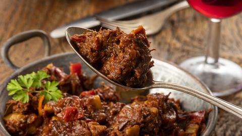 Gulaschportion auf Gabel. Rotweinglas und Brot im Hintergrund.