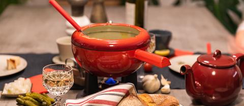 Ein Fondue-Topf auf einem Tisch, dazu diverse Beilagen (Brot, Gurke etc.)