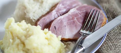 Auf einem Teller liegt eine Scheibe Kassler, dahinter Kartoffelbrei und Kraut, daneben Silberbesteck.
