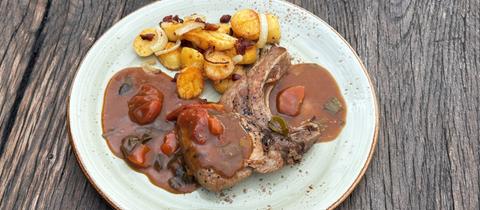 ein rohes Schweinekotelett auf einem Holzschneidebrett mit schwarzem Pfeffer, geschnittenen Zwiebeln und frischer grüner Petersilie.