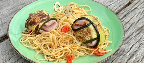 Gebackene Zucchini-Schinkenröllchen auf tomatisierten Spaghetti, serviert auf einem grünen Teller auf Holzgrund.