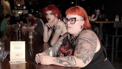 Zwei Protagonist:innen sitzen in einer Bar, im Hintergrund Menschen.