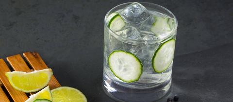 Mulled GinGin Tonic mit Gurke und auf Eis. Neben dem Cocktailglas liegen mehrere Limetten- und Gurkenscheiben, Zimtstangen, Gewürznelken, Kardamom und Wacholderbeeren.