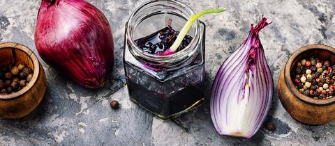 Rote-Zwiebel-Marmelade im Gläschen, mit aufgeschnittener roten Zwiebel und Döschen von Pfefferkörnern nebendran.