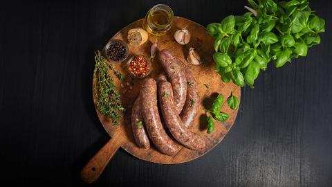 Bratwürste mit verschiedenen Gewürzen und Kräutern auf einem runden Holzbrett.