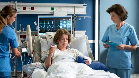 Viktoria Stadler (Isabella Krieger, l.) und Prof. Patzelt (Marijam Agischewa, r.) sind verwundert, dass ihre Patientin Beatrice Schneider (Laura Louisa Garde, M.), ihre erhebliche Verletzung als harmlos abtut.
