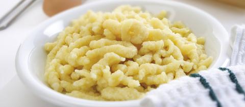 Nackte Spätzle in einem tiefen Teller, mit Küchenhandtuch, Mehl, Ei und Nudelholz im Hintergrund.