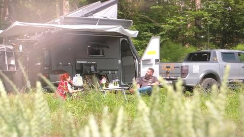 Protagonisten sitzen mit Camping-Fahrzeug und Zelt im Feld.
