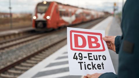 Jemand steht am Bahnhof und hält einen Zettel in der Hand, worauf steht: "DB 49 Euro Ticket"