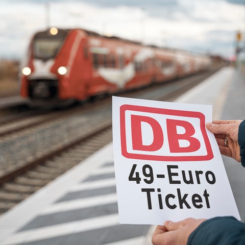 Jemand steht am Bahnhof und hält einen Zettel in der Hand, worauf steht: "DB 49 Euro Ticket"