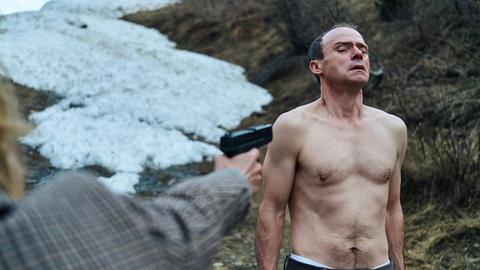 Ein Mann mit nacktem Oberkörper wird in den Bergen mit einer Waffe bedroht. 
