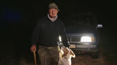 Ein Trüffeljäger mit Hund bei Dunkelheit vor den Scheinwerferlichtern seines Autos. 