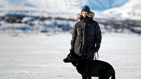 Frau mit schwarzer Winterjacke geht in Winterlandschaft mit Schäferhund spazieren. 