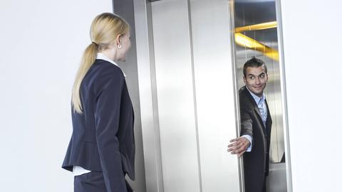 Ein Mann hält einer Frau die Aufzugstüre auf. 