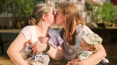 Zwei Frauen mit jeweils zwei Babys im Arm sitzen in einem Park und küssen sich.