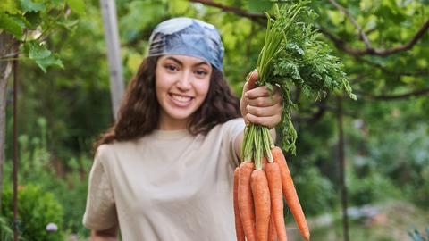 Eine junge Gärtnerin lächelnd mit einem Bund Karotten in der Hand. 