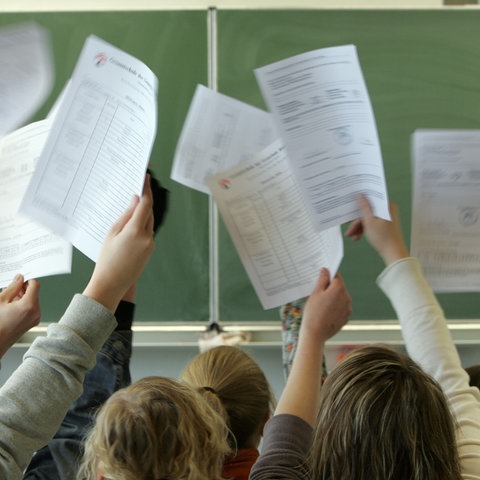 Schüler*innen halten vor der Tafel ihre Halbjahreszeugnisse nach oben in die Luft.