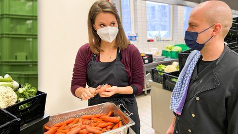 7 Tage-Reporterin steht mit einem Mann vor mehreren Gemüsekisten in einer Großküche und schält Karotten. Beide tragen einen Mundschutz.