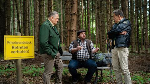 Zwei Polizisten unterhalten sich im Wald mit einem Mann, der auf einer Bank sitzt.