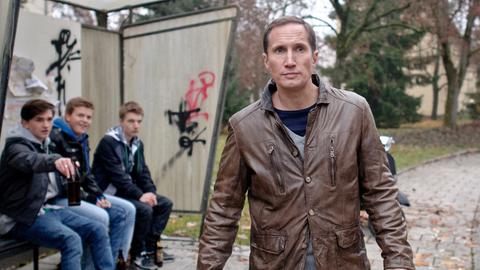 Ein Mann mit zwei Plastiktüten in den Händen läuft an einer Bushaltestelle vorbei, an der drei Jugendliche sitzen. Einer von ihnen zeigt auf den Mann.