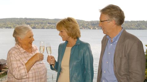 Zwei Frauen stoßen mit Sekt an. Ein Mann daneben schaut zu. Im Hintergrund ein See.