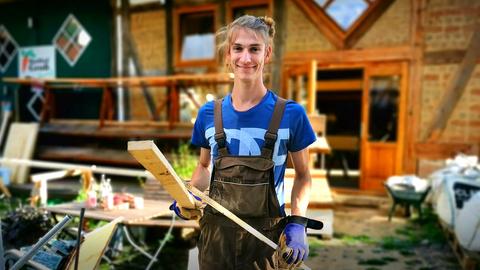 Protagonist mit Holzlatte vor Hofscheune und posiert für Foto.