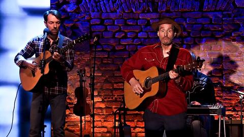 Dietrich Faber steht mit einer Gitarre auf der Bühne und schneidet eine Grimasse. Neben ihm steht ein weiterer Gitarrenspieler.