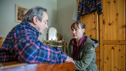 Eine Frau hockt vor einem älteren Mann, der im Sessel sitzt und traurig den Kopf hängen lässt.