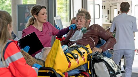 Marco Schiebel (Frederik Schmid, M.) hat bei dem Großunfall eine Fraktur und schwere Verbrennungen erlitten. Viktoria Stadler (Isabella Krieger, 2.v.r.) übernimmt den Patienten in der Notaufnahme von den Sanitäter:innen (Komparsen:innen).